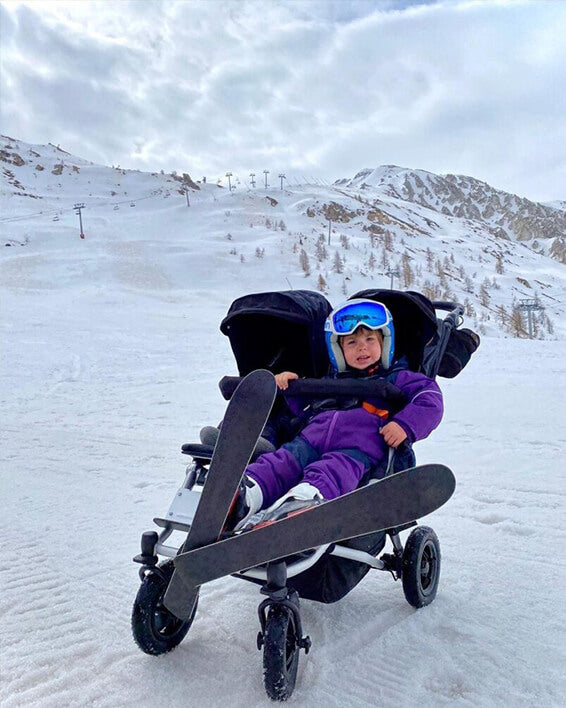child dressed in ski suit with both skis on sitting in duet™ four wheeled double buggy at skiing field - Mountain Buggy life without limit