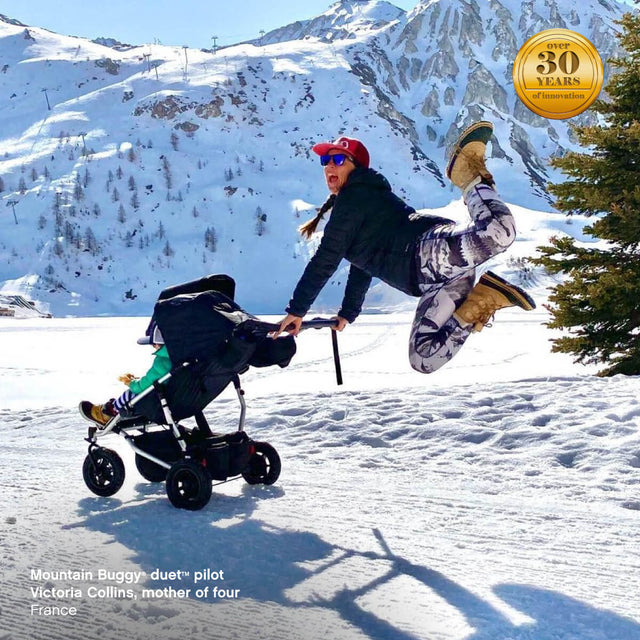 active mum pushing duet pram while jumping for joy at the snow field - Mountain Buggy duet™ pilot Victoria Collins, mother of four, France